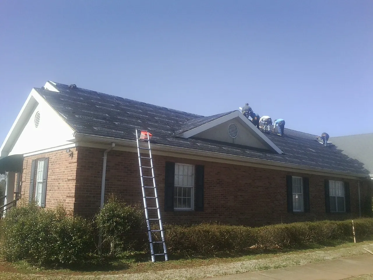 Crew tearing shingles off a brick ranch roof with a ladder set — Emergency Roof Repair work by our crew in Grainfield