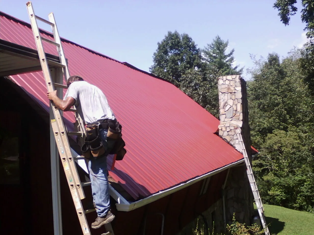 Roofer on a ladder beside a red metal roof and stone chimney — Soffit & Fascia Repair by Roofing Brothers in Grainfield